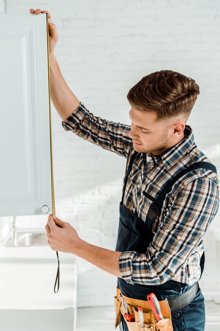handsome installer in overalls measuring white kitchen cabinet door